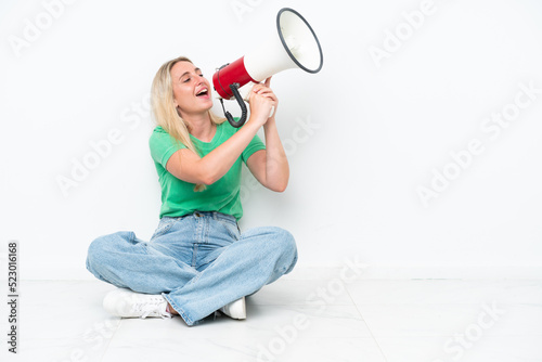 Young caucasian woman sitting on the floor isolated on white background shouting through a megaphone