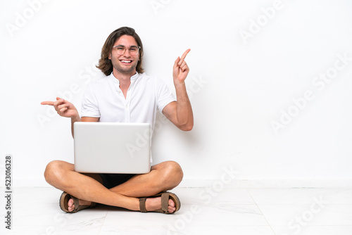 Young handsome man with a laptop sitting on the floor isolated on white background pointing finger to the laterals and happy © luismolinero