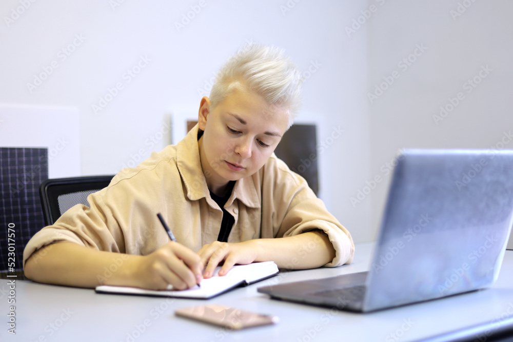 Girl with short blonde hair writes in a notebook while sitting at the desk with laptop. Tomboy lifestyle, organization of office work and planning Girl with short blonde hair writes in a notebook while sitting at the desk with laptop. Tomboy lifestyle, organization of office work and planning