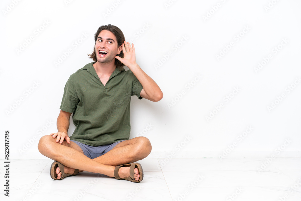 Young handsome man sitting on the floor isolated on white background listening to something by putting hand on the ear Young handsome man sitting on the floor isolated on white background listening to something by putting hand on the ear
