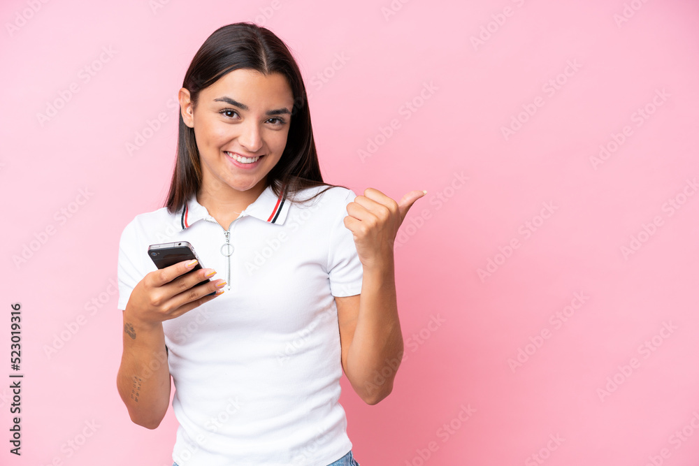 Young caucasian woman isolated on pink background using mobile phone and pointing to the lateral Young caucasian woman isolated on pink background using mobile phone and pointing to the lateral