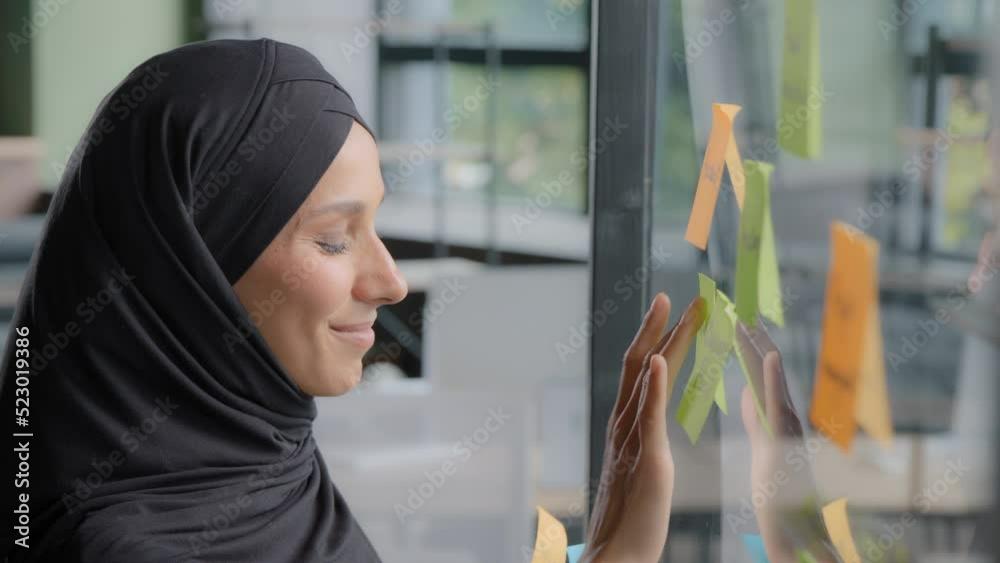 Smiling successful businesswoman corporate leader writing project tasks leaves reminder on sticky colored note organizes work young arab woman office worker in hijab attaches stickers on glass board