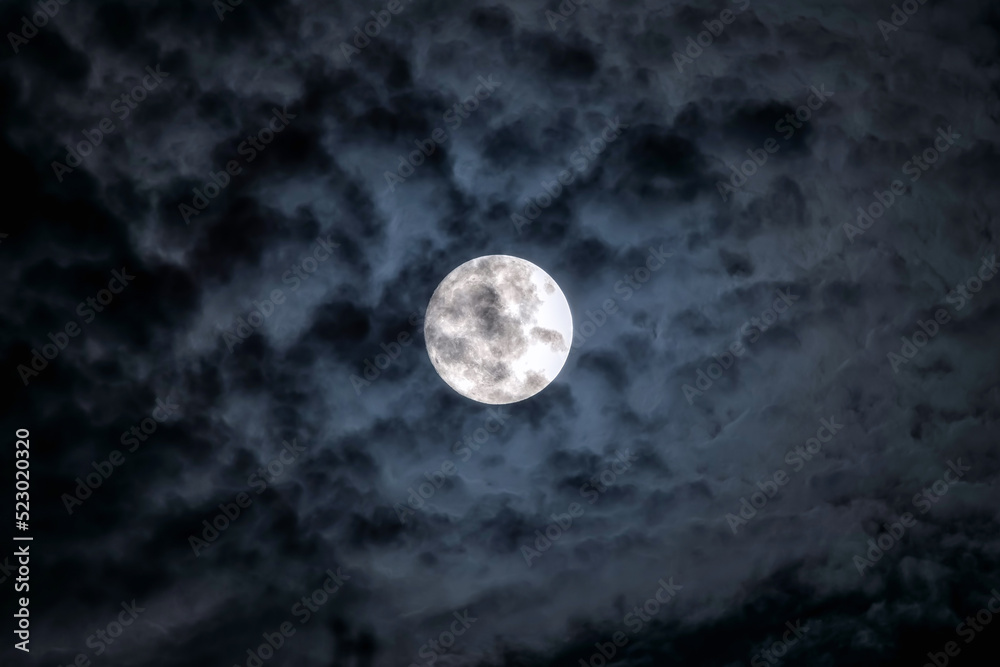 Full moon at night. The illuminated face of the moon is wrapped in a cloud cover that covers it in a veiled way. Full moon at night. The illuminated face of the moon is wrapped in a cloud cover that covers it in a veiled way.