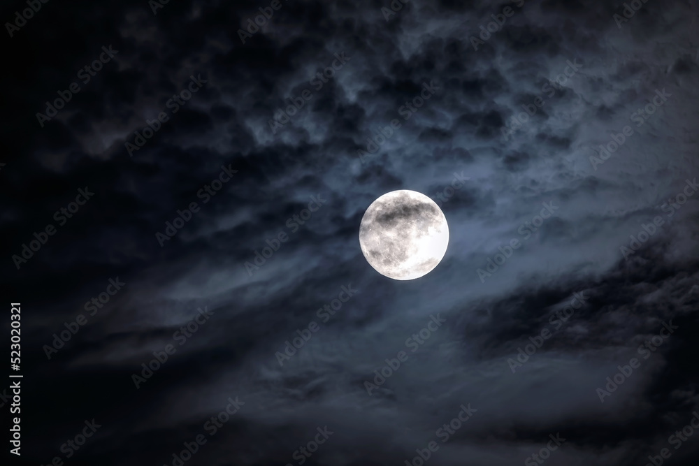 Full moon at night. The illuminated face of the moon is wrapped in a cloud cover that covers it in a veiled way. Full moon at night. The illuminated face of the moon is wrapped in a cloud cover that covers it in a veiled way.