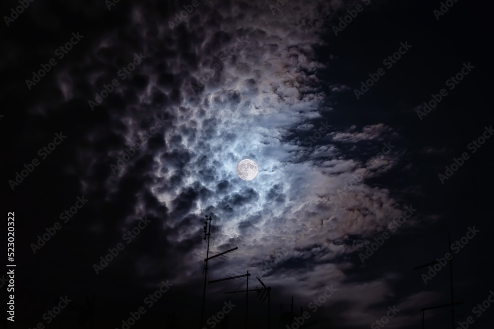 Full moon at night. The illuminated face of the moon is wrapped in a cloud cover that covers it in a veiled way. Full moon at night. The illuminated face of the moon is wrapped in a cloud cover that covers it in a veiled way.