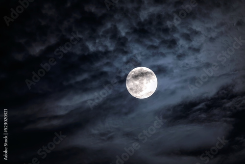 Full moon at night. The illuminated face of the moon is wrapped in a cloud cover that covers it in a veiled way.