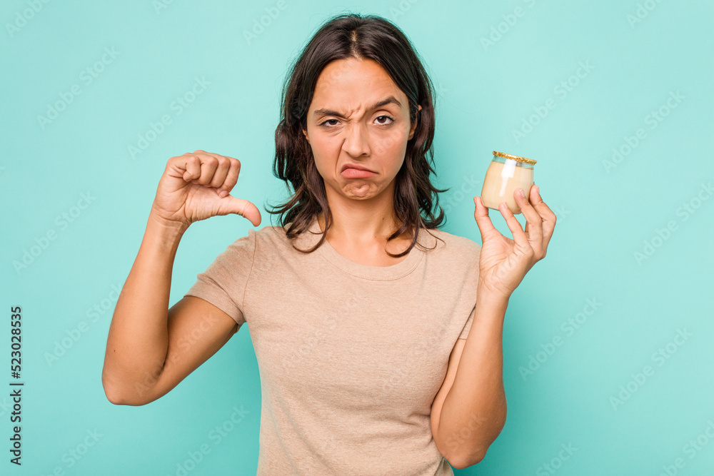 Young hispanic woman holding yogurt isolated on blue background showing a dislike gesture, thumbs down. Disagreement concept. Young hispanic woman holding yogurt isolated on blue background showing a dislike gesture, thumbs down. Disagreement concept.