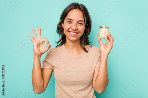 Young hispanic woman holding yogurt isolated on blue background cheerful and confident showing ok gesture.