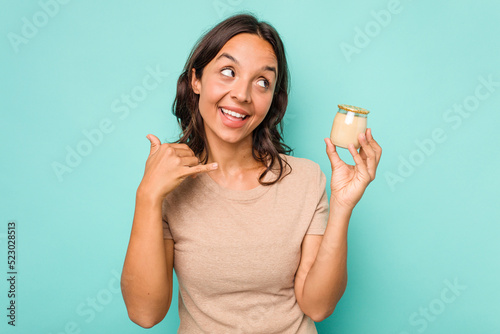 Young hispanic woman holding yogurt isolated on blue background showing a mobile phone call gesture with fingers.