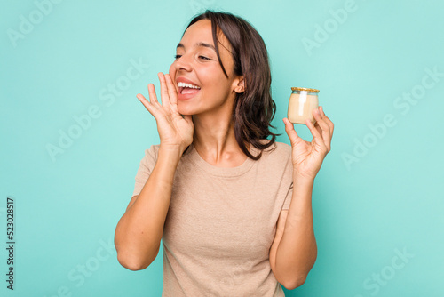 Young hispanic woman holding yogurt isolated on blue background shouting and holding palm near opened mouth.
