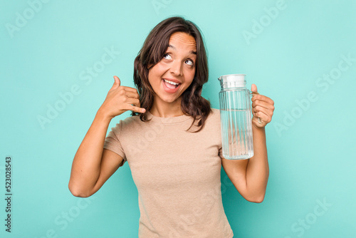 Young hispanic woman holding a water of jar isolated on blue background showing a mobile phone call gesture with fingers.