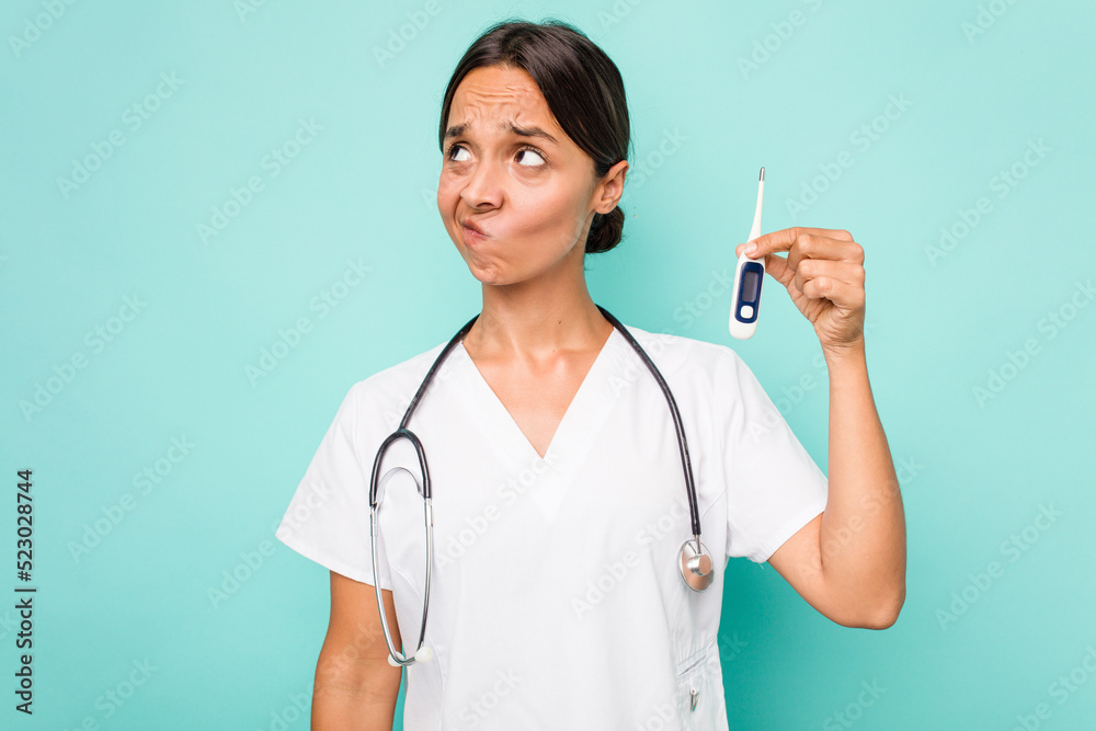 Young hispanic nurse woman holding a thermometer isolated on blue background confused, feels doubtful and unsure. Young hispanic nurse woman holding a thermometer isolated on blue background confused, feels doubtful and unsure.