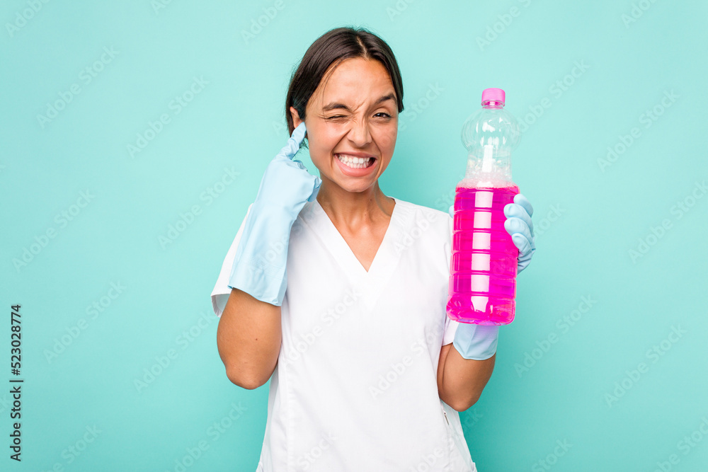 Young cleaner hispanic woman isolated on blue background covering ears with hands. Young cleaner hispanic woman isolated on blue background covering ears with hands.