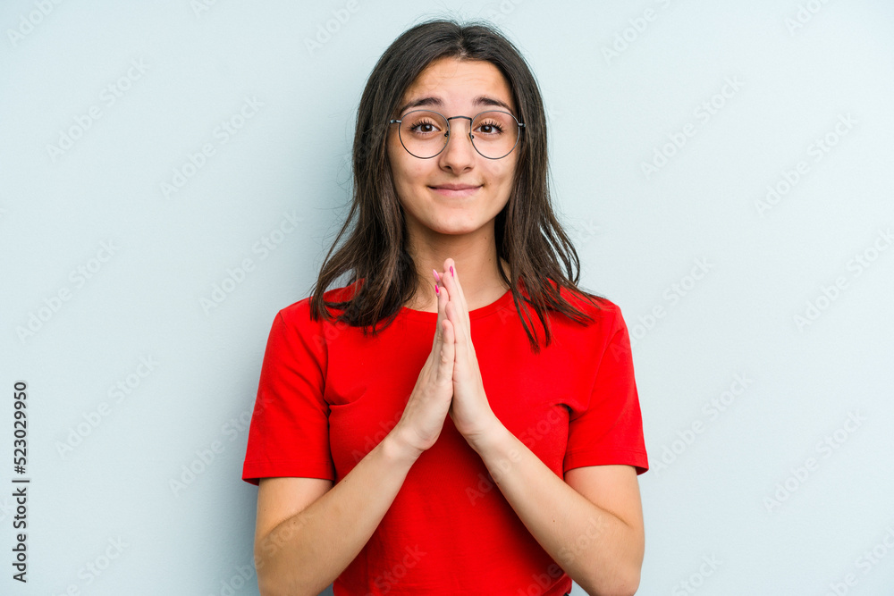 Fototapeta premium Young caucasian woman isolated on blue background holding hands in pray near mouth, feels confident.