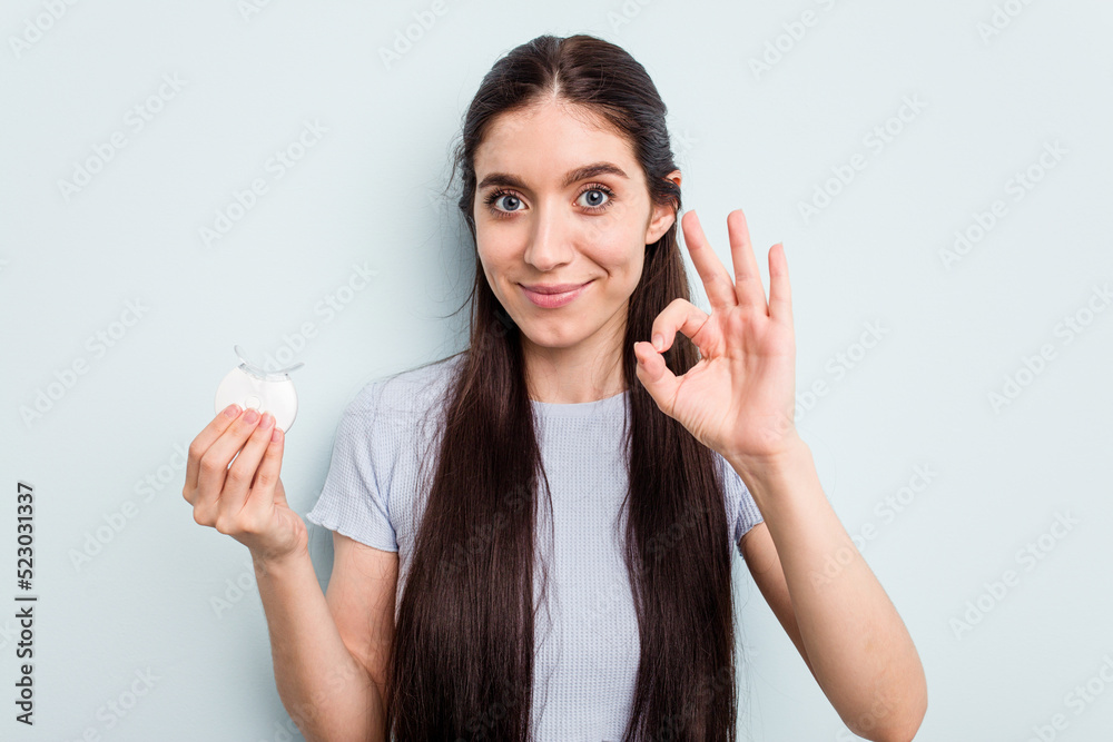 Young caucasian woman holding teeth whitener isolated on blue background cheerful and confident showing ok gesture. Young caucasian woman holding teeth whitener isolated on blue background cheerful and confident showing ok gesture.