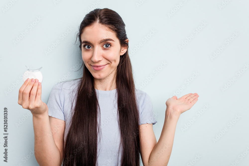 Young caucasian woman holding teeth whitener isolated on blue background showing a copy space on a palm and holding another hand on waist. Young caucasian woman holding teeth whitener isolated on blue background showing a copy space on a palm and holding another hand on waist.