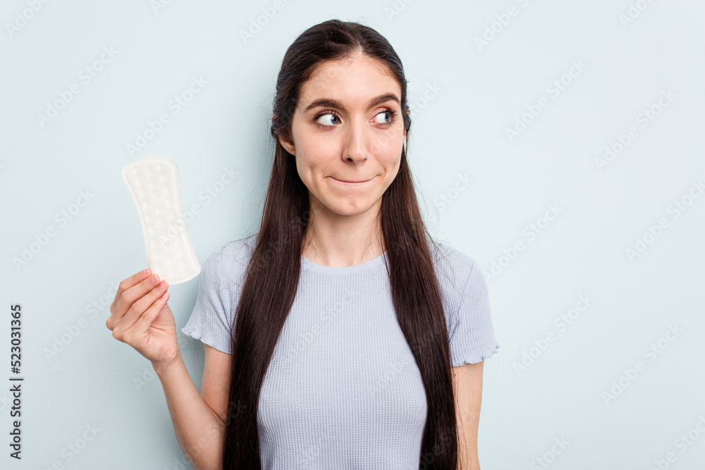 Young caucasian woman holding a sanitary napkin isolated on blue background confused, feels doubtful and unsure. Young caucasian woman holding a sanitary napkin isolated on blue background confused, feels doubtful and unsure.