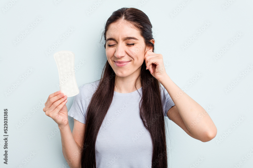 Young caucasian woman holding a sanitary napkin isolated on blue background covering ears with hands. Young caucasian woman holding a sanitary napkin isolated on blue background covering ears with hands.
