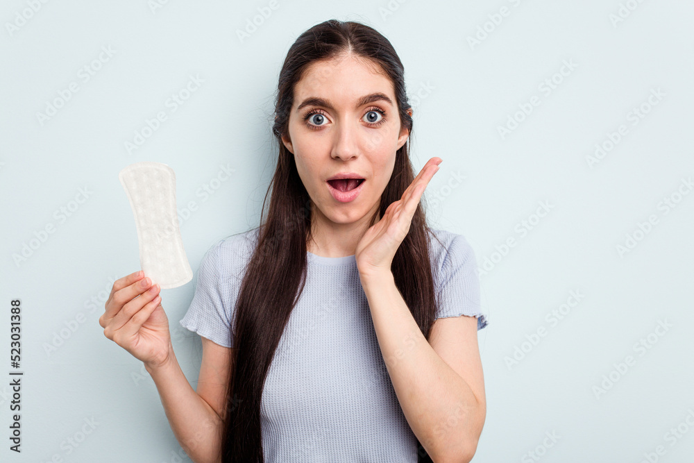 Young caucasian woman holding a sanitary napkin isolated on blue background surprised and shocked. Young caucasian woman holding a sanitary napkin isolated on blue background surprised and shocked.