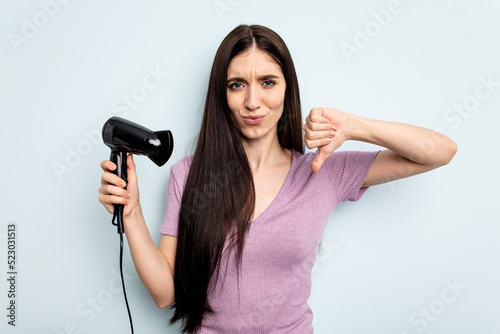 Young caucasian woman holding hairdryer isolated on blue background showing a dislike gesture, thumbs down. Disagreement concept.