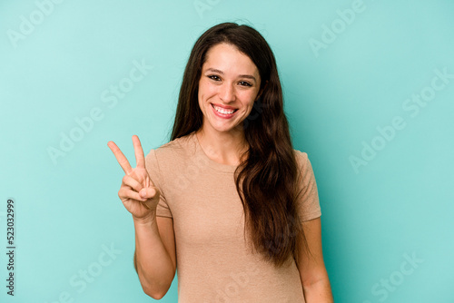 Young caucasian woman isolated on blue background showing victory sign and smiling broadly.