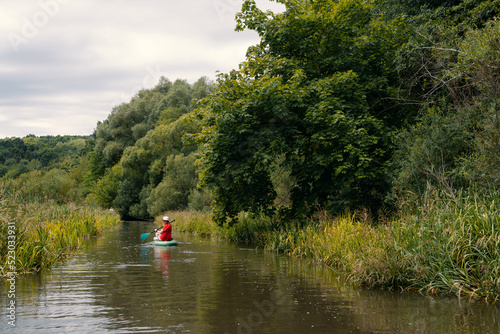 A narrow river among thickets of bushes.. Girl surfer in red t-shirt sitting on the sup board. Paddleboard SUP Water Sport Activity Holidays Vacation. Beautiful nature outdoors. Copy space.