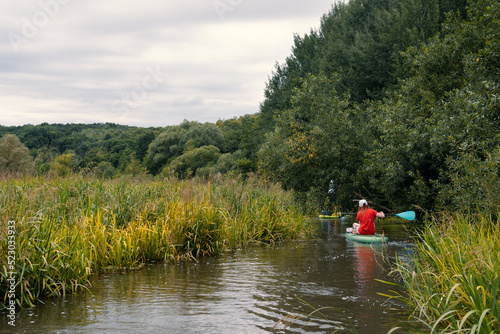 A narrow river among thickets of bushes.. Girl surfer in red t-shirt sitting on the sup board. Paddleboard SUP Water Sport Activity Holidays Vacation. Beautiful nature outdoors. Copy space.