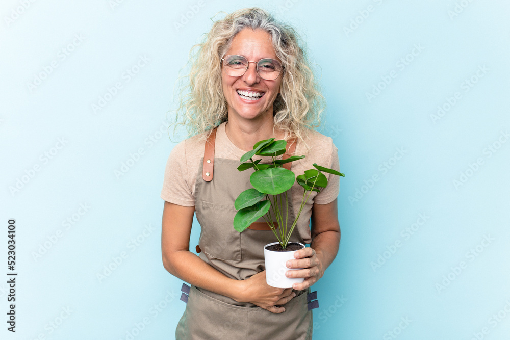 Middle age gardener woman holding a plant isolated on blue background laughing and having fun. Middle age gardener woman holding a plant isolated on blue background laughing and having fun.
