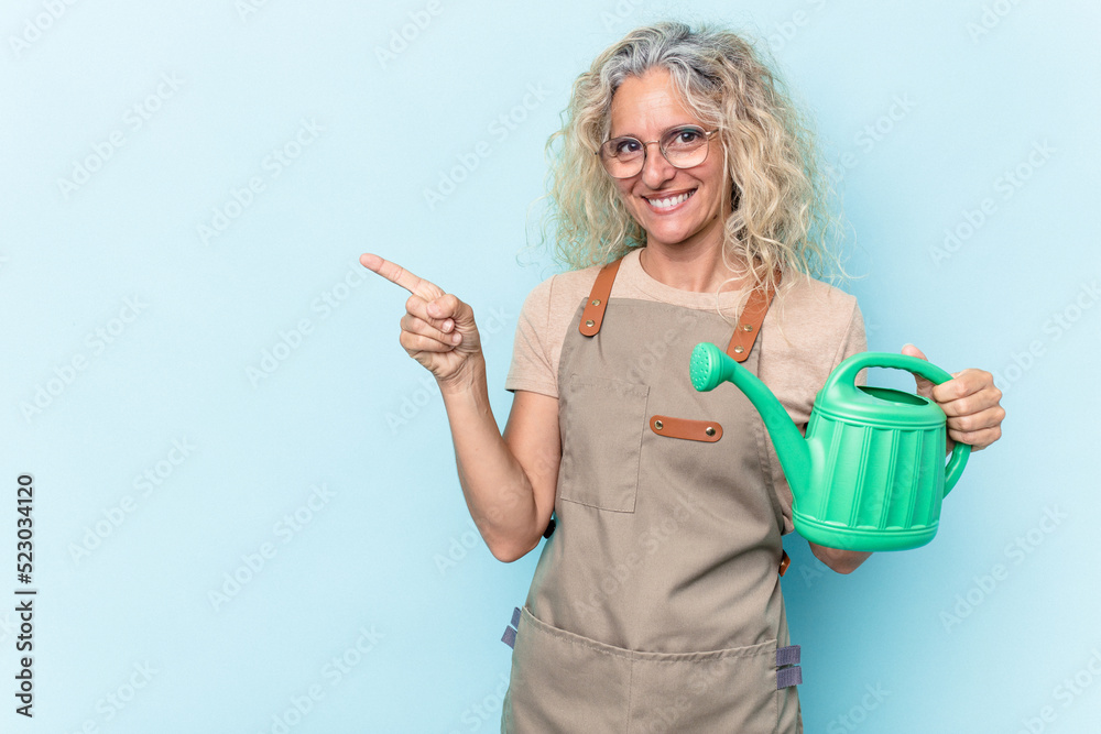 Middle age gardener woman holding a watering can isolated on white background smiling and pointing aside, showing something at blank space.