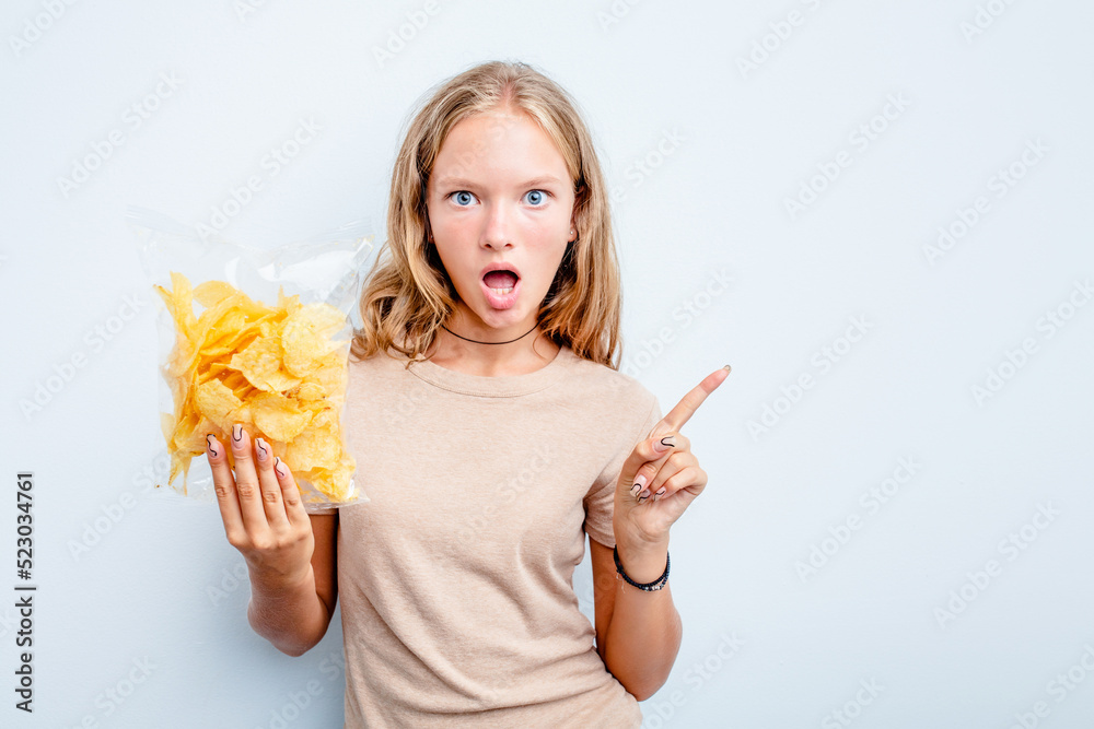 Caucasian teen girl holding bag of chips isolated on blue background pointing to the side Caucasian teen girl holding bag of chips isolated on blue background pointing to the side