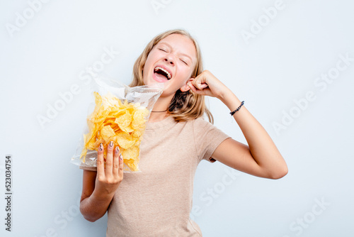 Caucasian teen girl holding bag of chips isolated on blue background raising fist after a victory  winner concept.