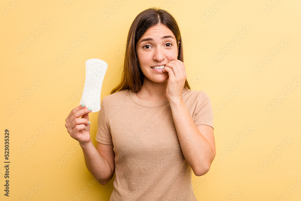 Young caucasian woman holding a sanitary napkin isolated on yellow background biting fingernails, nervous and very anxious. Young caucasian woman holding a sanitary napkin isolated on yellow background biting fingernails, nervous and very anxious.