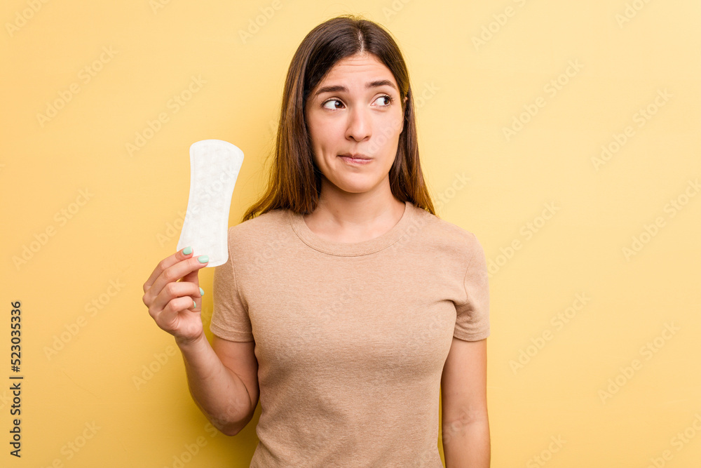 Young caucasian woman holding a sanitary napkin isolated on yellow background confused, feels doubtful and unsure. Young caucasian woman holding a sanitary napkin isolated on yellow background confused, feels doubtful and unsure.