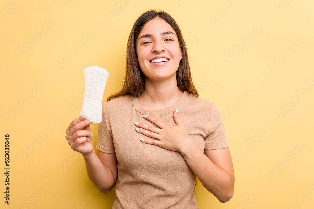 Young caucasian woman holding a sanitary napkin isolated on yellow background laughs out loudly keeping hand on chest. Young caucasian woman holding a sanitary napkin isolated on yellow background laughs out loudly keeping hand on chest.