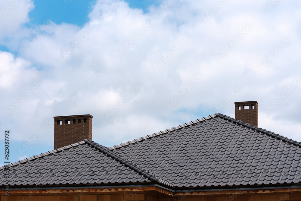 Dark metal tile shingles roof with chimney against blue sky with white clouds. Dark metal tile shingles roof with chimney against blue sky with white clouds.