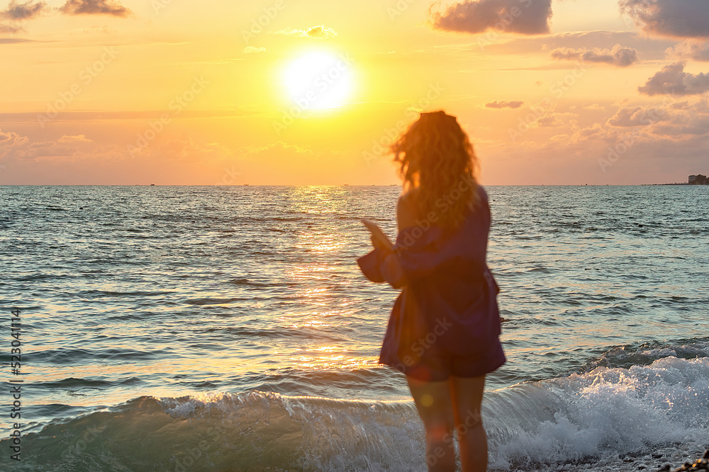silhouette of a girl with long wavy hair out of focus, she spends summer time in the evening at sunset on the seashore, in her hands is a tablet, selective focus silhouette of a girl with long wavy hair out of focus, she spends summer time in the evening at sunset on the seashore, in her hands is a tablet, selective focus