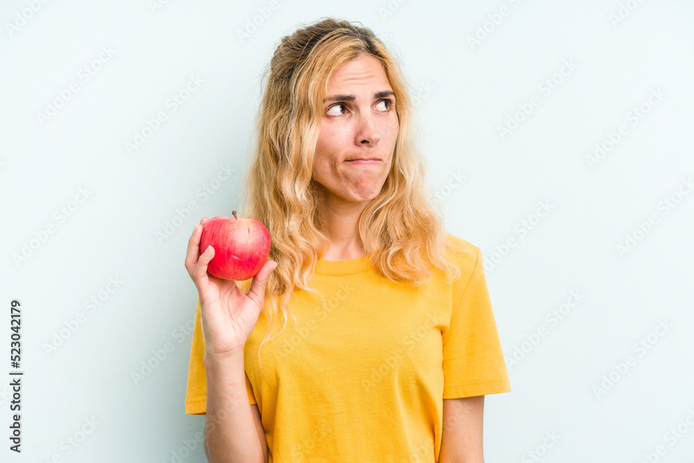 Young caucasian woman holding an apple isolated on blue background confused, feels doubtful and unsure. Young caucasian woman holding an apple isolated on blue background confused, feels doubtful and unsure.