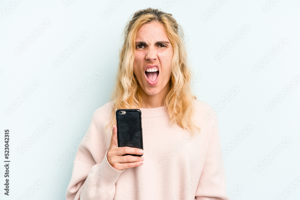 Young caucasian woman holding mobile phone isolated on blue background screaming very angry and aggressive. Young caucasian woman holding mobile phone isolated on blue background screaming very angry and aggressive.