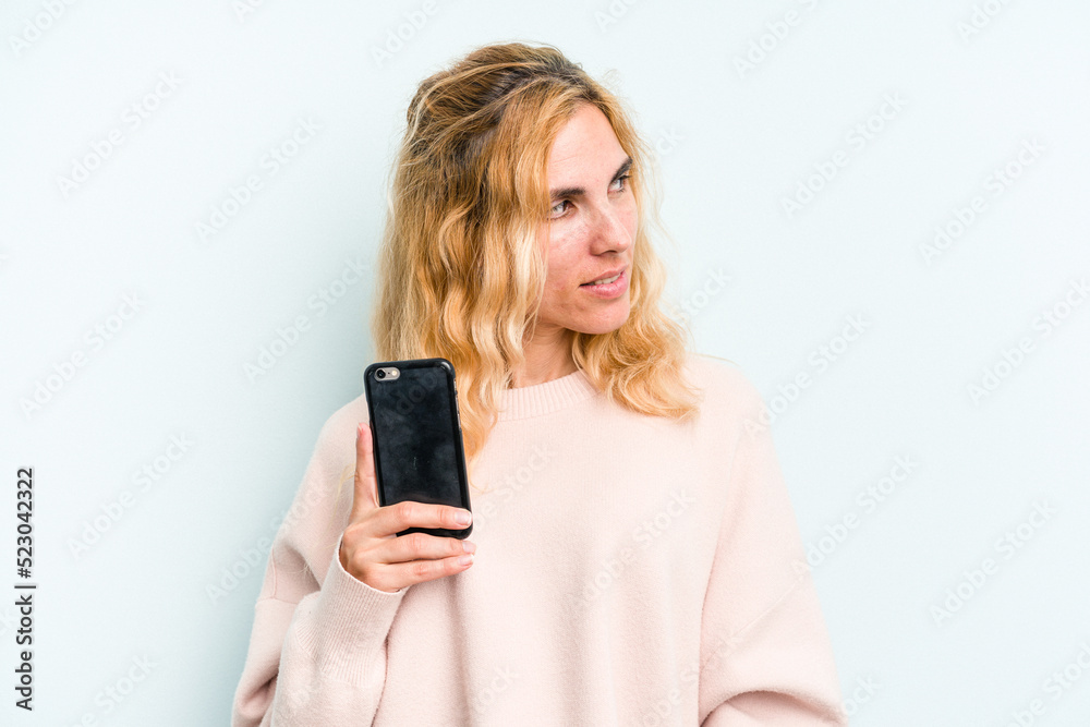 Young caucasian woman holding mobile phone isolated on blue background looks aside smiling, cheerful and pleasant. Young caucasian woman holding mobile phone isolated on blue background looks aside smiling, cheerful and pleasant.