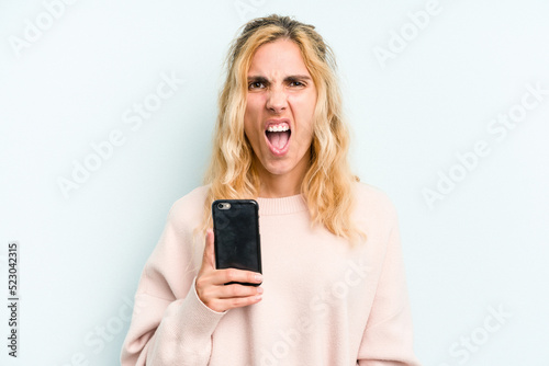 Young caucasian woman holding mobile phone isolated on blue background screaming very angry and aggressive.