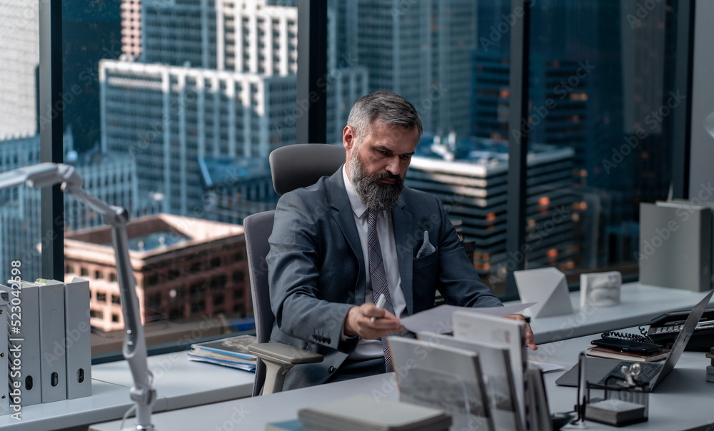 Portrait of 50s adult grey-haired Caucasian CEO businessman working at his desk in the office located in a skyscraper, business district buildings in the background Portrait of 50s adult grey-haired Caucasian CEO businessman working at his desk in the office located in a skyscraper, business district buildings in the background
