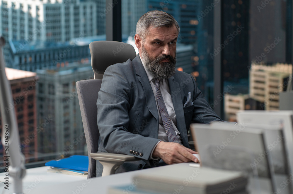 Portrait of 50s adult grey-haired Caucasian CEO businessman working at his desk in the office located in a skyscraper, business district buildings in the background Portrait of 50s adult grey-haired Caucasian CEO businessman working at his desk in the office located in a skyscraper, business district buildings in the background
