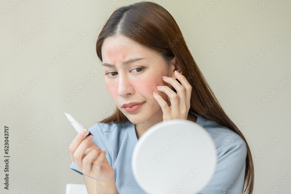 Dermatology, asian young woman looking at mirror, holding cream tube in hand, expression worry and itch, itchy allergy or allergic sensitive reaction, red or rash on face. Beauty care of skin problem Dermatology, asian young woman looking at mirror, holding cream tube in hand, expression worry and itch, itchy allergy or allergic sensitive reaction, red or rash on face. Beauty care of skin problem
