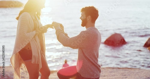 Romantic man proposing to woman on beach at sunset and hugging, embracing or holding his girlfriend after she says yes. Happy, smiling or excited couple getting engaged with a ring near sea or ocean photo