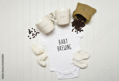 Baby Announcement. Baby Brewing written on a onesie with booties Coffee mugs and Spilled Coffee Beans. Flat lay on white beadboard with copy space.