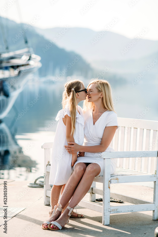Happy millenial mother and blonde teenager daughter in white family look dresses sits near seashore  in a touristic  luxury resort with mountains  and yachts