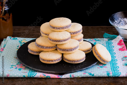 Torre de alfajores de maicena sobre plato de pizarra. Dulce tradicional argentino. Sándwich de galletas relleno de dulce de leche con coco rallado. Fondo negro. 