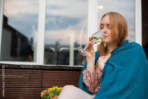 Fotografie Portrait of young buxom woman covered by blue blanket sitting near cut fruits on tray, drinking glass of white wine