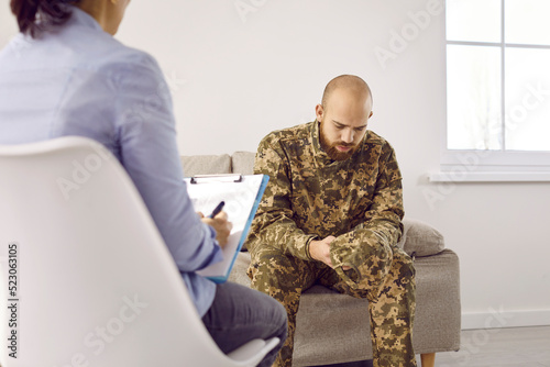 Depressed soldier who's gone through horrors of war and is suffering from military PTSD is sharing his concerns and worries at therapy session with professional psychologist who's writing on clipboard