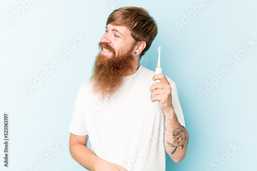 Young caucasian man holding a electric toothbrush isolated on blue background looks aside smiling, cheerful and pleasant.